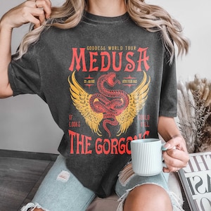 K&ouml;nnte beinhalten: Ein schwarzes T-Shirt mit einem Grafikdruck einer rot-gelben gefl&uuml;gelten Schlange mit dem Text "GODDESS WORLD TOUR MEDUSA 22:00 HR 9TH FEB 1962 IF LOOKS THE GORGO COULD MILL"