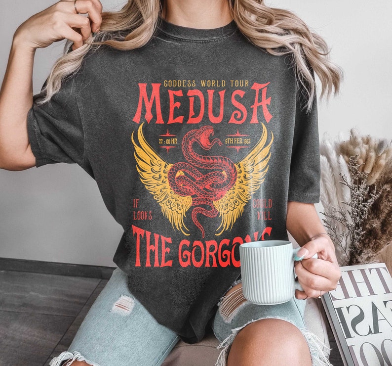 K&ouml;nnte beinhalten: Ein schwarzes T-Shirt mit einem Grafikdruck einer rot-gelben gefl&uuml;gelten Schlange mit dem Text "GODDESS WORLD TOUR MEDUSA 22:00 HR 9TH FEB 1962 IF LOOKS THE GORGO COULD MILL"