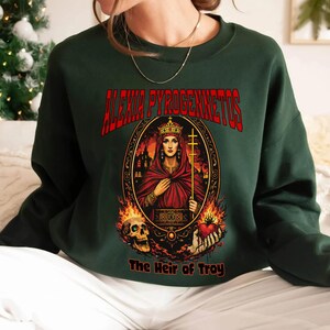 Op de afbeelding: Donkergroene sweatshirt met een afbeelding van een gekroonde vrouw in een rode mantel, met een kruis en een hart. De tekst "ALEXIA PYROGENNETOS" staat in het rood boven de afbeelding, met "The Heir of Troy" eronder.