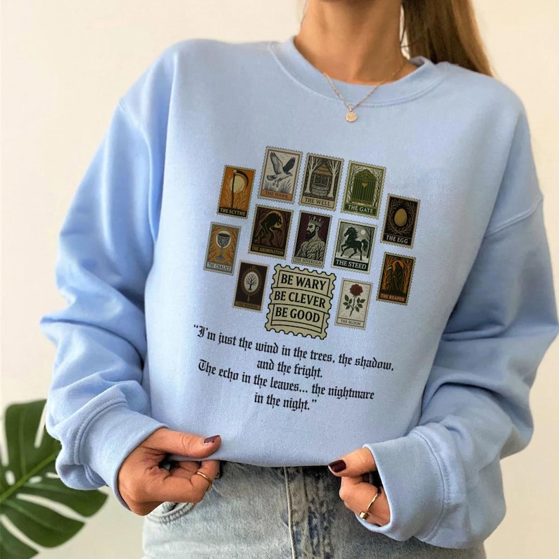 May include: Light blue sweatshirt with a collection of vintage-style postage stamp graphics. The stamps feature various illustrations and the phrases "BE WARY BE CLEVER BE GOOD" and a quote about the wind and trees.