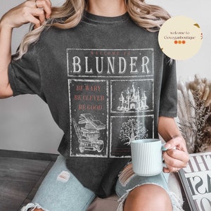 Op de afbeelding: Een zwart T-shirt met een wit grafisch ontwerp met de tekst "Welkom bij Blunder" en illustraties van een kasteel, een stapel boeken en een boom. De tekst "Wees voorzichtig, Wees slim, Wees goed" is ook in het ontwerp opgenomen.