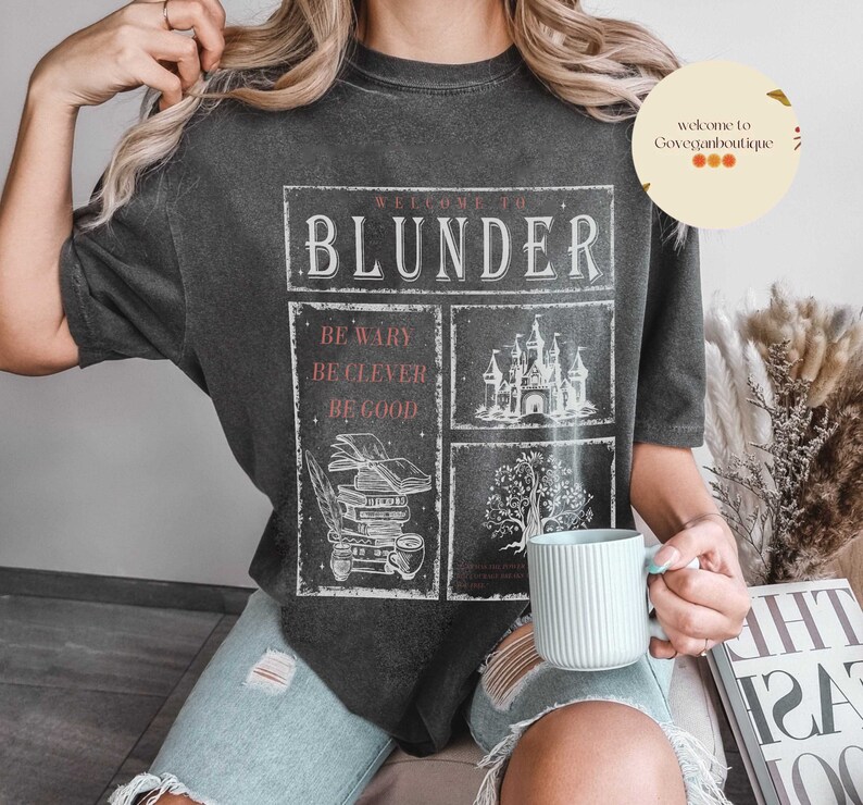 Op de afbeelding: Een zwart T-shirt met een wit grafisch ontwerp met de tekst "Welkom bij Blunder" en illustraties van een kasteel, een stapel boeken en een boom. De tekst "Wees voorzichtig, Wees slim, Wees goed" is ook in het ontwerp opgenomen.