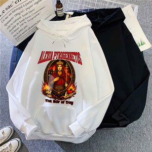 Op de afbeelding: Witte hoodie met een afbeelding van een gekroonde vrouw, vlammen en de tekst "ALEXIA PYROGENNETOS" en "The Heir of Troy". Een zwarte hoodie ligt ernaast.