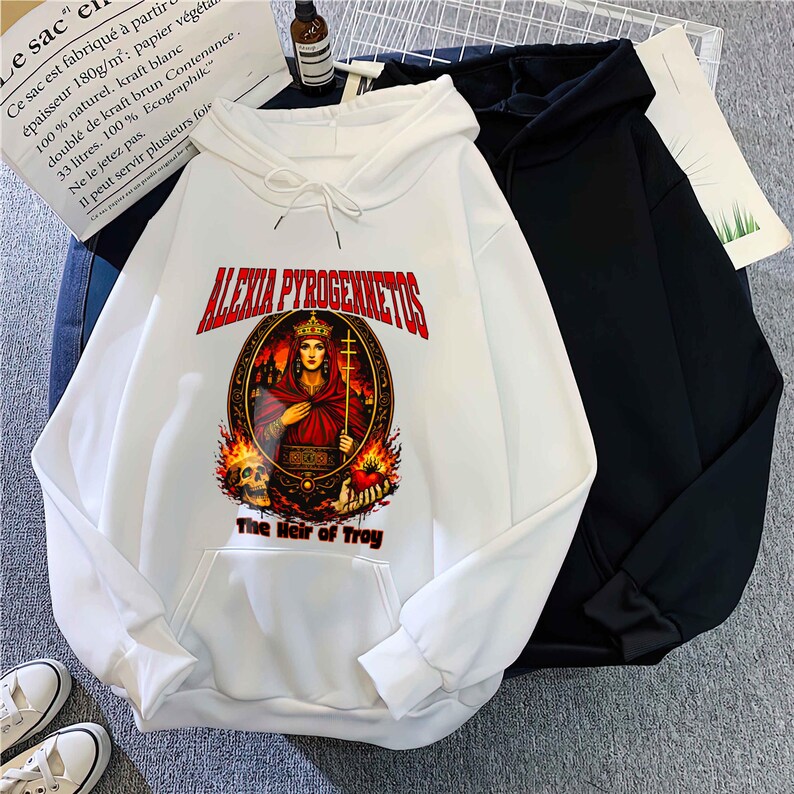 Op de afbeelding: Witte hoodie met een afbeelding van een gekroonde vrouw, vlammen en de tekst "ALEXIA PYROGENNETOS" en "The Heir of Troy". Een zwarte hoodie ligt ernaast.