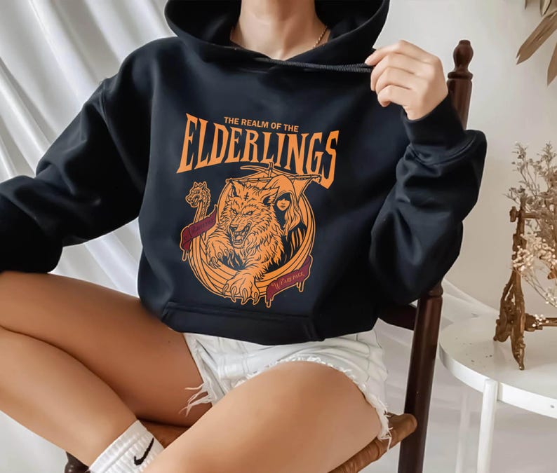 May include: Black hooded sweatshirt featuring an orange graphic with a wolf, a woman, and the text "THE REALM OF THE ELDERLINGS". The design is encircled by a maroon banner with text. The sweatshirt has a drawstring hood.