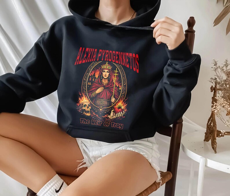 Op de afbeelding: Zwarte hoodie met de rode tekst "ALEXIA PYROGENNETOS" en een illustratie van een gekroonde vrouw die een kruis vasthoudt. Daaronder staat "The Heir of Troy". Het ontwerp bevat vlammen en een schedel.