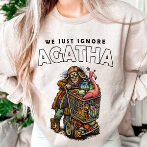 May include: Beige sweatshirt with the text "WE JUST IGNORE AGATHA". The graphic shows a person pushing a shopping cart filled with items, including an IKEA box and a pink flamingo. The person is wearing a hat and glasses.
