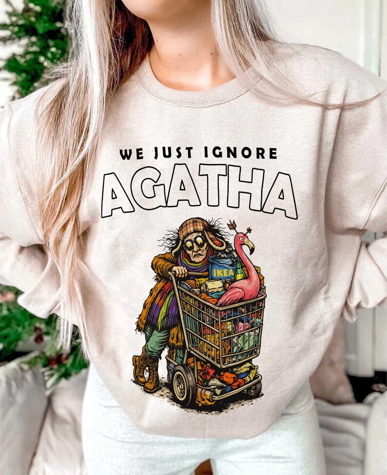 May include: Beige sweatshirt with the text "WE JUST IGNORE AGATHA". The graphic shows a person pushing a shopping cart filled with items, including an IKEA box and a pink flamingo. The person is wearing a hat and glasses.