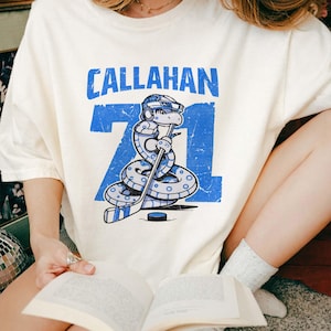 May include: Cream-colored t-shirt featuring a blue graphic of a snake wearing a hockey helmet and holding a hockey stick. The text "CALLAHAN 71" is printed above the snake. The design has a distressed, vintage look.