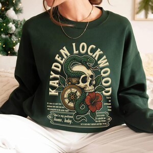 May include: Dark green sweatshirt featuring a graphic design with the text "KAYDEN LOCKWOOD". The design includes a skull, snake, compass, and a red hibiscus flower. The sweatshirt has long sleeves.
