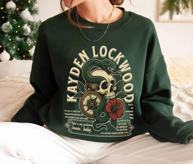 May include: Dark green sweatshirt featuring a graphic design with the text "KAYDEN LOCKWOOD". The design includes a skull, snake, compass, and a red hibiscus flower. The sweatshirt has long sleeves.