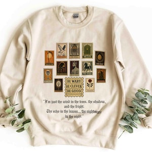 May include: Cream-colored sweatshirt featuring a collection of postage stamp-style illustrations with text. The stamps depict various images, including a scythe, a well, and a gate. The phrase "BE WARY BE CLEVER BE GOOD" is also printed on the sweatshirt.