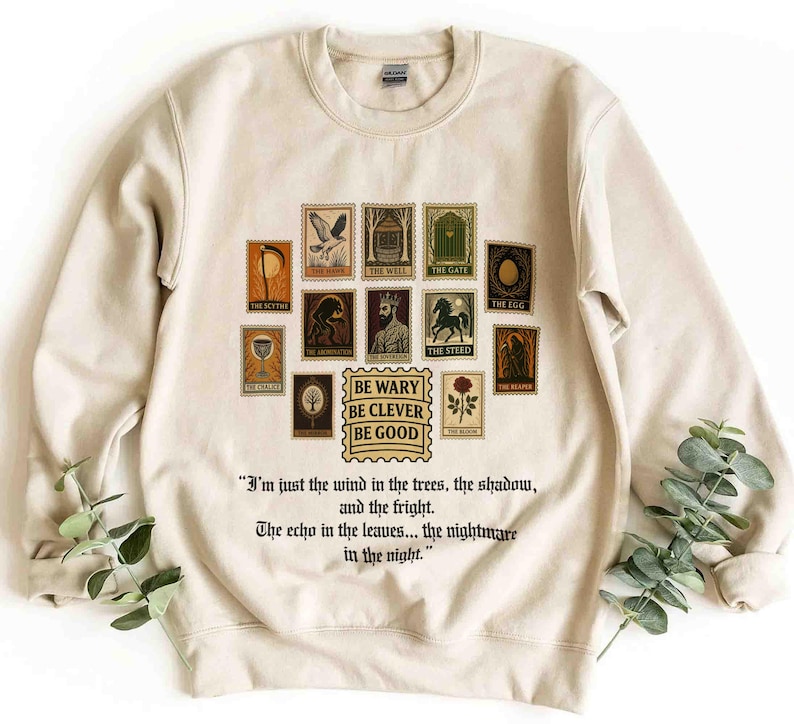 May include: Cream-colored sweatshirt featuring a collection of postage stamp-style illustrations with text. The stamps depict various images, including a scythe, a well, and a gate. The phrase "BE WARY BE CLEVER BE GOOD" is also printed on the sweatshirt.