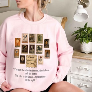 May include: A light pink sweatshirt featuring a collection of vintage-style postage stamps with various illustrations. Below the stamps is the text "BE WARY BE CLEVER BE GOOD" and a quote. The sweatshirt is a comfortable, casual garment.