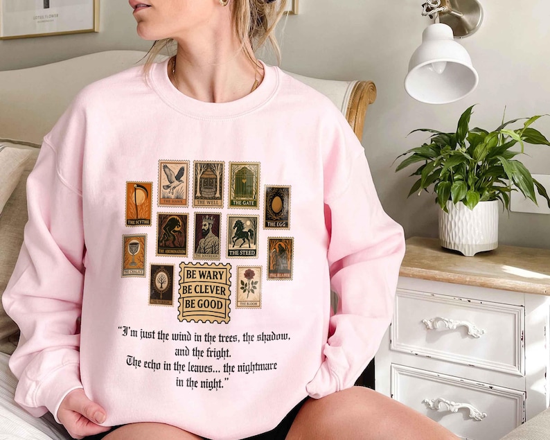 May include: A light pink sweatshirt featuring a collection of vintage-style postage stamps with various illustrations. Below the stamps is the text "BE WARY BE CLEVER BE GOOD" and a quote. The sweatshirt is a comfortable, casual garment.