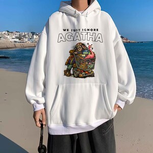 May include: White hoodie with a front graphic featuring a person pushing a shopping cart filled with items, including a pink flamingo. The text "WE JUST IGNORE AGATHA" is above the graphic. The hoodie has a kangaroo pocket and a drawstring hood.