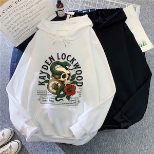 May include: White hoodie with a front graphic featuring a skull, snake, compass, and red flower, with the text "KAYDEN LOCKWOOD". A black hoodie is next to it. The white hoodie has a front pocket and drawstring hood.