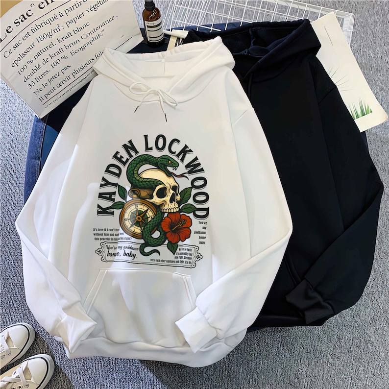 May include: White hoodie with a front graphic featuring a skull, snake, compass, and red flower, with the text "KAYDEN LOCKWOOD". A black hoodie is next to it. The white hoodie has a front pocket and drawstring hood.