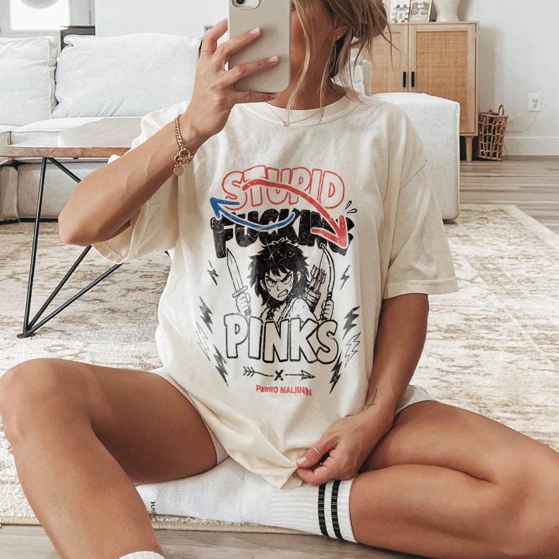 May include: Cream-colored t-shirt with a graphic design featuring the words "STUPID FUCKING PINKS" in red, blue, and black. The design includes an illustration of a character holding weapons. The text "PERRO MALINN" is below the graphic.