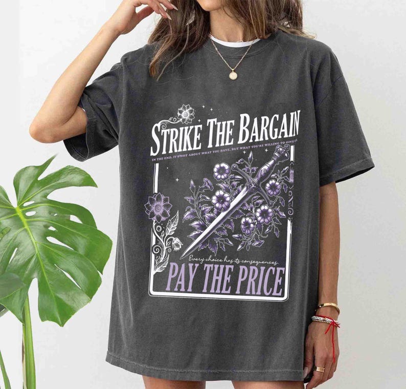 May include: A gray oversized t-shirt with a graphic print of a sword and flowers. The text on the graphic reads "Strike the Bargain" and "Pay the Price".
