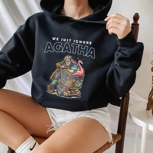May include: Black hooded sweatshirt with the text "WE JUST IGNORE AGATHA" in white. The graphic depicts a person pushing a shopping cart filled with items, including a pink flamingo. The cart has the IKEA logo.