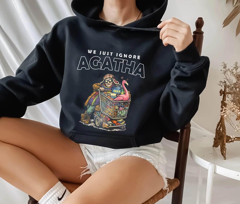 May include: Black hooded sweatshirt with the text "WE JUST IGNORE AGATHA" in white. The graphic depicts a person pushing a shopping cart filled with items, including a pink flamingo. The cart has the IKEA logo.