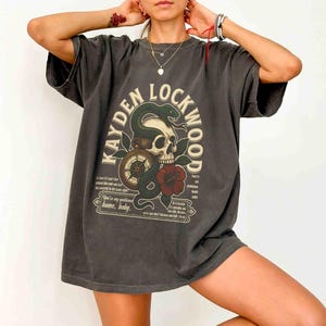 May include: Dark gray t-shirt featuring the text "KAYDEN LOCKWOOD" in an arch above a skull, snake, compass, and red flower design. The shirt has additional text below the graphic. The shirt is oversized.