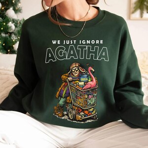 May include: Dark green sweatshirt with the text "WE JUST IGNORE AGATHA". The graphic depicts a person pushing a shopping cart filled with items, including a pink flamingo. The cart has the IKEA logo.