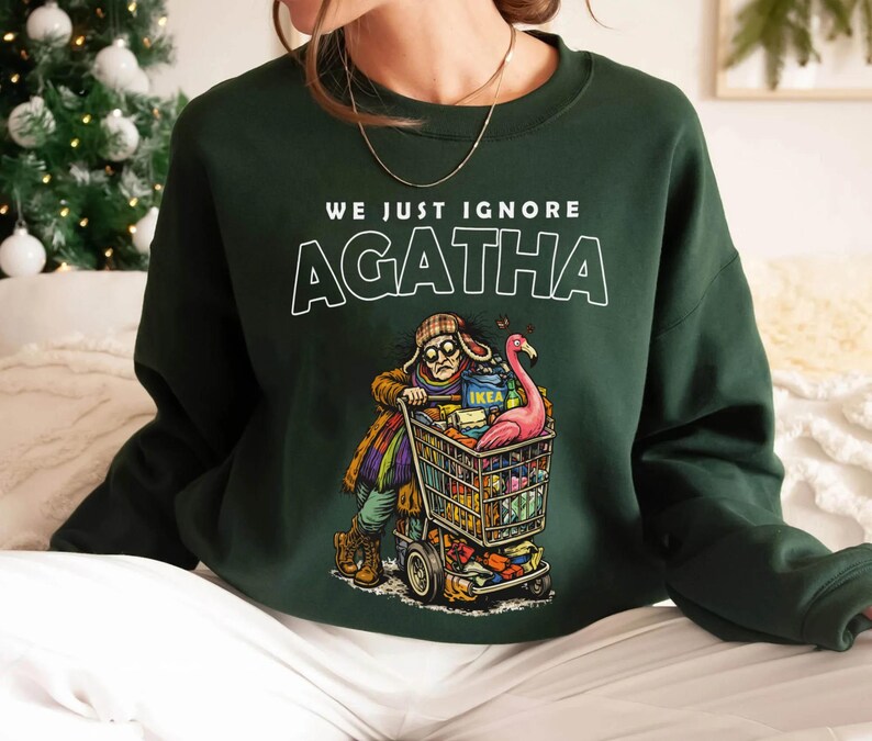May include: Dark green sweatshirt with the text "WE JUST IGNORE AGATHA". The graphic depicts a person pushing a shopping cart filled with items, including a pink flamingo. The cart has the IKEA logo.