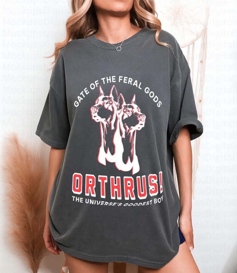 Puede incluir: Camiseta gris oscuro con un gr&aacute;fico de un perro de dos cabezas y el texto "GATE OF THE FERAL GODS" arriba y "ORTHRUS! THE UNIVERSE'S GOODEST BOY" abajo. El dise&ntilde;o es en rojo y blanco.