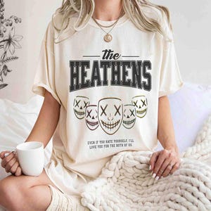 May include: Cream-colored t-shirt featuring the text "The Heathens" in a bold, black, collegiate-style font. Below the text are five stylized mask graphics with an "X" for eyes and a stitched smile. The shirt also has the quote: "EVEN IF YOU HATE YOURSELF, I'LL LOVE YOU FOR THE BOTH OF US."