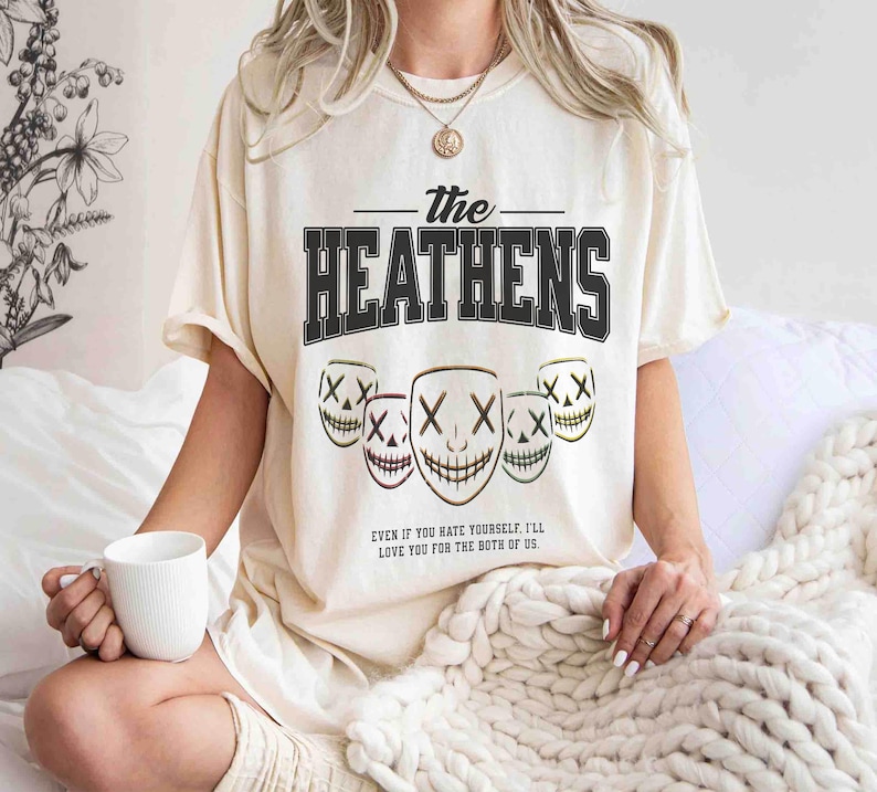 May include: Cream-colored t-shirt featuring the text "The Heathens" in a bold, black, collegiate-style font. Below the text are five stylized mask graphics with an "X" for eyes and a stitched smile. The shirt also has the quote: "EVEN IF YOU HATE YOURSELF, I'LL LOVE YOU FOR THE BOTH OF US."