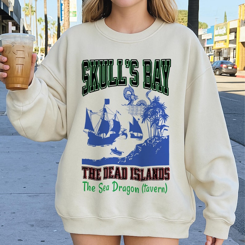 May include: Cream-colored sweatshirt featuring a pirate-themed graphic. The design includes the text "SKULL'S BAY" in green, a blue illustration of ships, islands, and a sea dragon, and the text "THE DEAD ISLANDS The Sea Dragon (tavern)" in red and green.