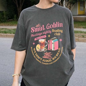 May include: Dark gray t-shirt with a graphic that reads "Smut Goblin meetings nightly Reading club." The design includes a spice level, a drink, books, and the text "We don't judge, we read."