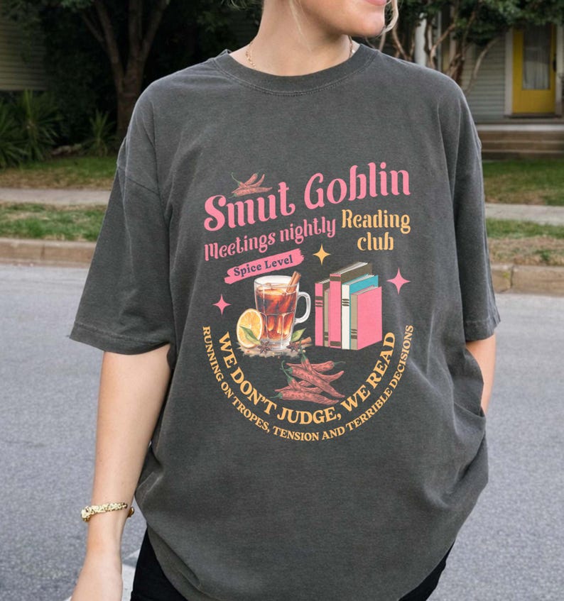 May include: Dark gray t-shirt with a graphic that reads "Smut Goblin meetings nightly Reading club." The design includes a spice level, a drink, books, and the text "We don't judge, we read."