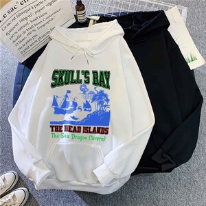 May include: White hooded sweatshirt with a front graphic that reads "SKULL'S BAY" above a pirate ship and island scene. The text "THE DEAD ISLANDS The Sea Dragon (tavern)" is below. A black hoodie is next to it.