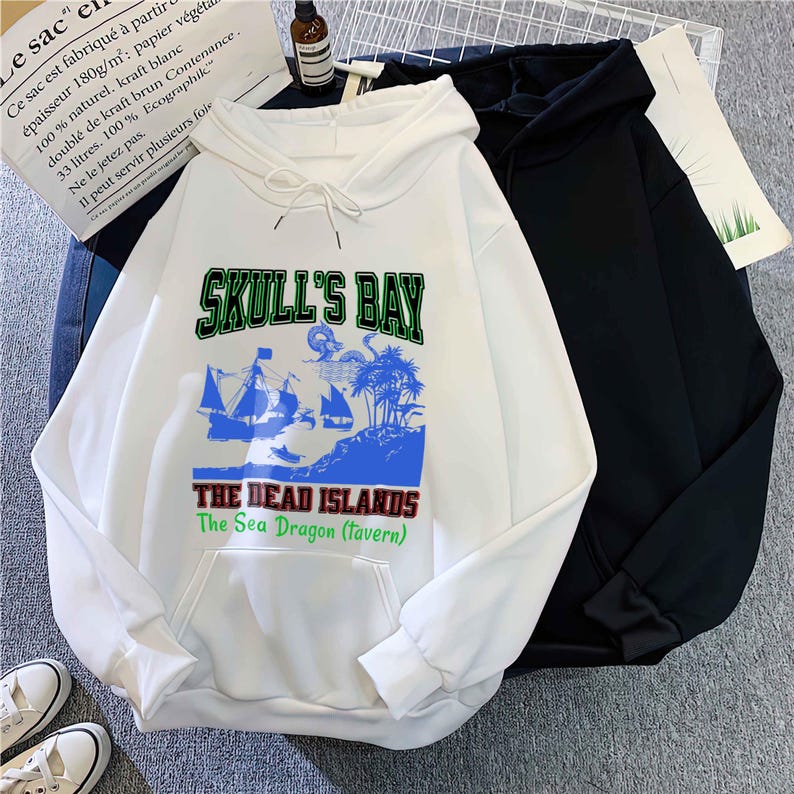 May include: White hooded sweatshirt with a front graphic that reads "SKULL'S BAY" above a pirate ship and island scene. The text "THE DEAD ISLANDS The Sea Dragon (tavern)" is below. A black hoodie is next to it.