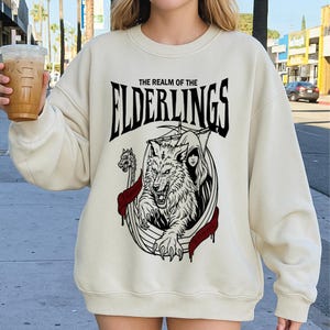 May include: Cream-colored sweatshirt with a black and white graphic. The graphic features a wolf, a woman, and the text "THE REALM OF THE ELDERLINGS". The design includes red accents and a Viking ship.