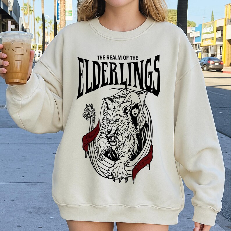 May include: Cream-colored sweatshirt with a black and white graphic. The graphic features a wolf, a woman, and the text "THE REALM OF THE ELDERLINGS". The design includes red accents and a Viking ship.