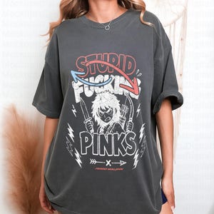 May include: Dark grey t-shirt with a graphic design. The design features the words "STUPID FUCKING PINKS" in red and white, with an illustration of a character holding a knife and bow and arrow. The text "FERRO MALIGN" is below the graphic.