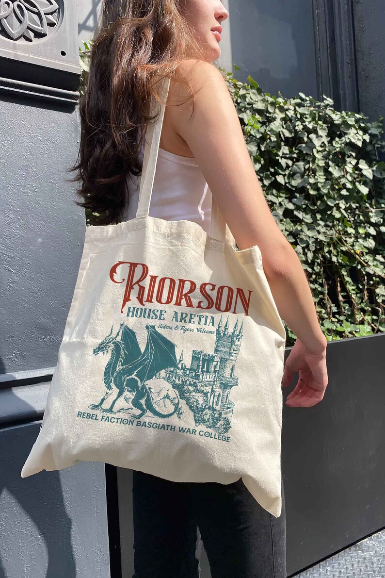 May include: A white canvas tote bag featuring a green dragon and castle graphic. The text on the bag reads "Riorson House Aretia Riders & Ryers Welcome Rebel Faction Basgiath War College".