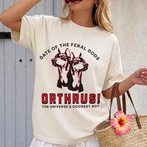Puede incluir: Camiseta color crema con un gr&aacute;fico de un perro de dos cabezas, el texto "GATE OF THE FERAL GODS" y "ORTHRUS! THE UNIVERSE'S GOODEST BOY" en rojo y negro. La camiseta tiene mangas cortas y un corte holgado.