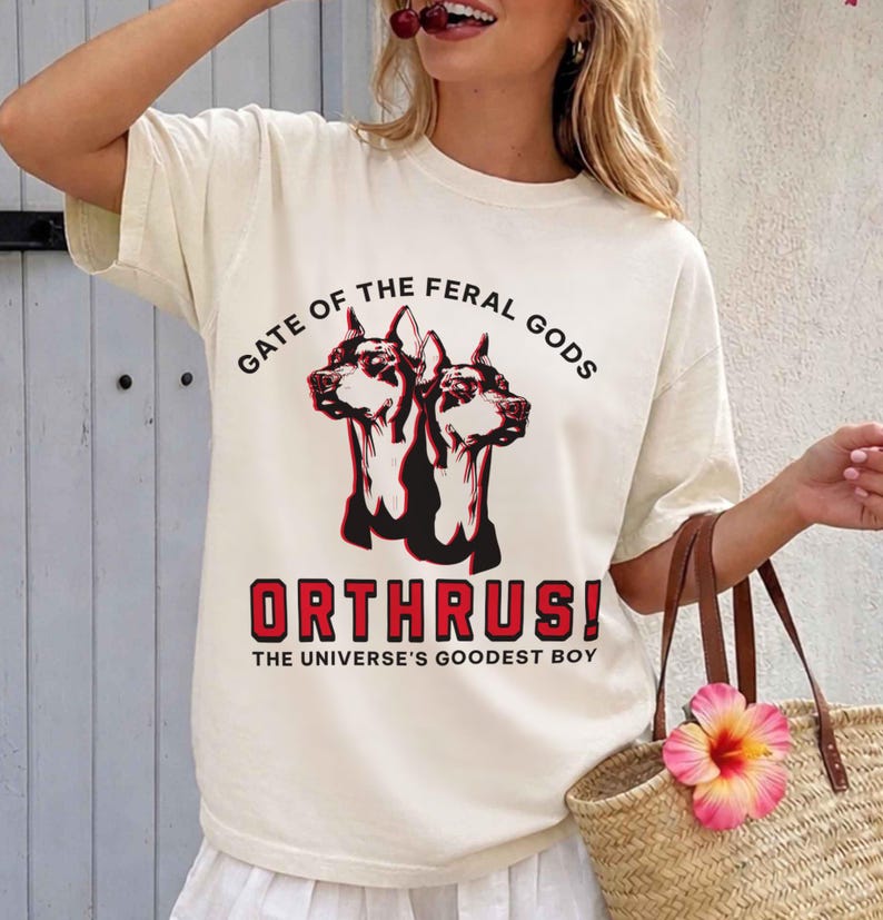Puede incluir: Camiseta color crema con un gr&aacute;fico de un perro de dos cabezas, el texto "GATE OF THE FERAL GODS" y "ORTHRUS! THE UNIVERSE'S GOODEST BOY" en rojo y negro. La camiseta tiene mangas cortas y un corte holgado.