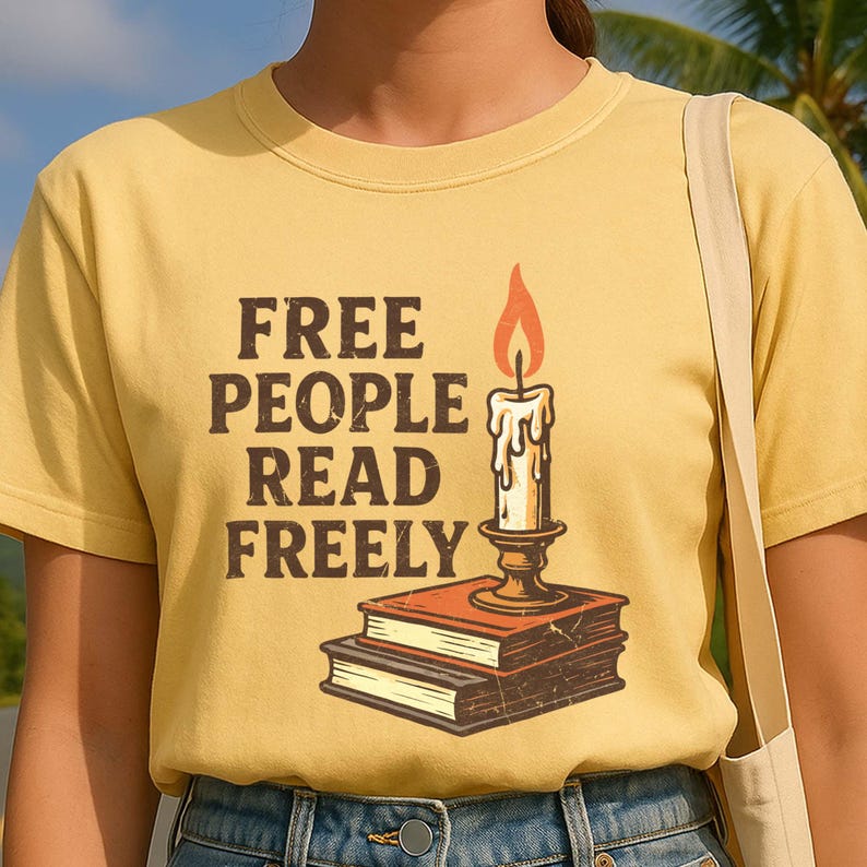 Może przedstawiać: Musztardowa koszulka z napisem "FREE PEOPLE READ FREELY" w brązowych literach. Świeca z płomieniem spoczywa na dw&oacute;ch książkach w grafice w stylu vintage.
