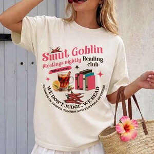 May include: Cream-colored t-shirt with a graphic design. The design features text that reads "Smut Goblin Reading Club" with illustrations of books, chili peppers, and a drink. The shirt has short sleeves.