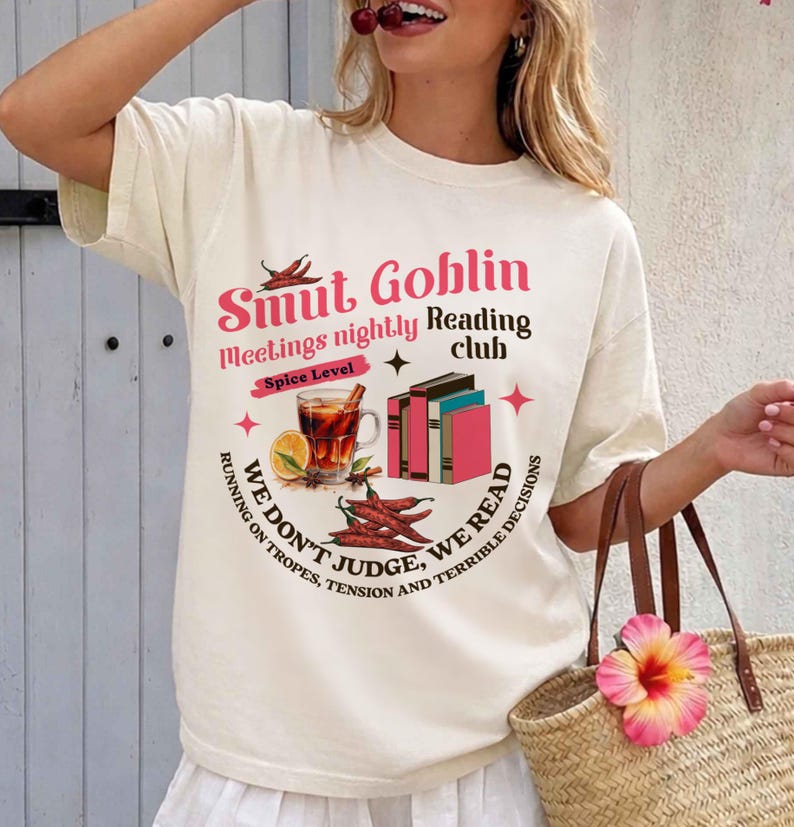 May include: Cream-colored t-shirt with a graphic design. The design features text that reads "Smut Goblin Reading Club" with illustrations of books, chili peppers, and a drink. The shirt has short sleeves.