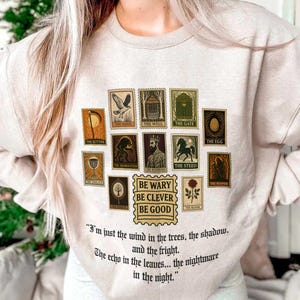 May include: Beige sweatshirt featuring a collection of illustrated stamps with various images and text. The stamps include images of a scythe, hawk, well, gate, and more. Below the stamps is the text "BE WARY BE CLEVER BE GOOD" and a quote.