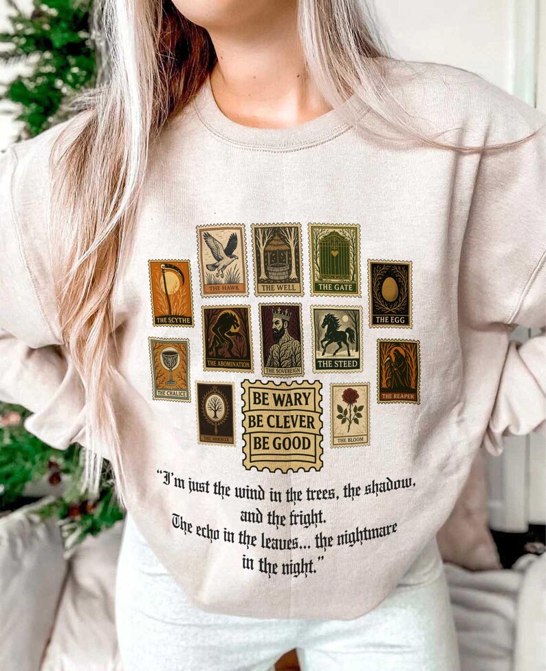 May include: Beige sweatshirt featuring a collection of illustrated stamps with various images and text. The stamps include images of a scythe, hawk, well, gate, and more. Below the stamps is the text "BE WARY BE CLEVER BE GOOD" and a quote.