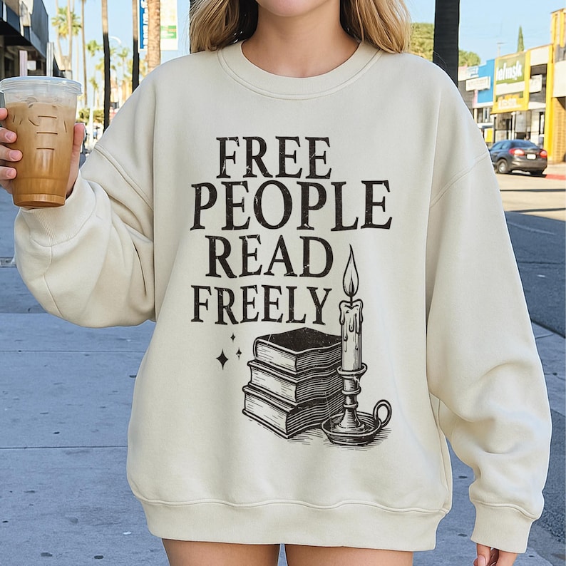 Op de afbeelding: Cr&egrave;mekleurige sweatshirt met de zwarte tekst "FREE PEOPLE READ FREELY". Daaronder een afbeelding van gestapelde boeken, een kaars en sterren. De sweatshirt heeft een casual stijl.