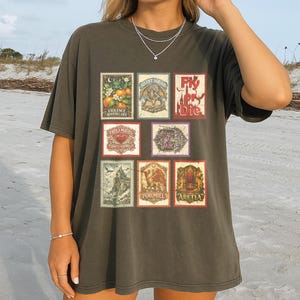 May include: Dark olive green t-shirt featuring a collection of vintage-style postage stamp designs. The stamps showcase various illustrations, including botanical elements, dragons, and text such as "Fly or Die" and "Violence Apothecary".